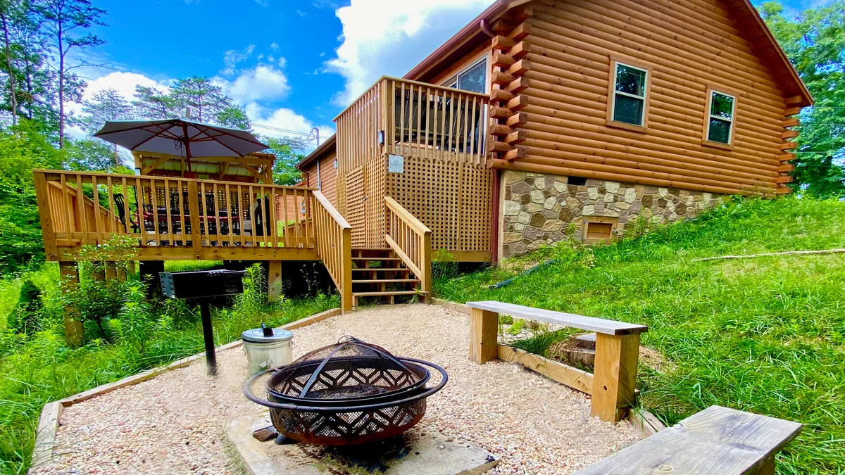 The image showcases a charming log cabin nestled in the woods, featuring a spacious wooden deck overlooking the trees, a fire pit surrounded by chairs, and a grill—perfect for outdoor gatherings. The sunset casts a warm glow over the scene, evoking feelings of relaxation and togetherness.