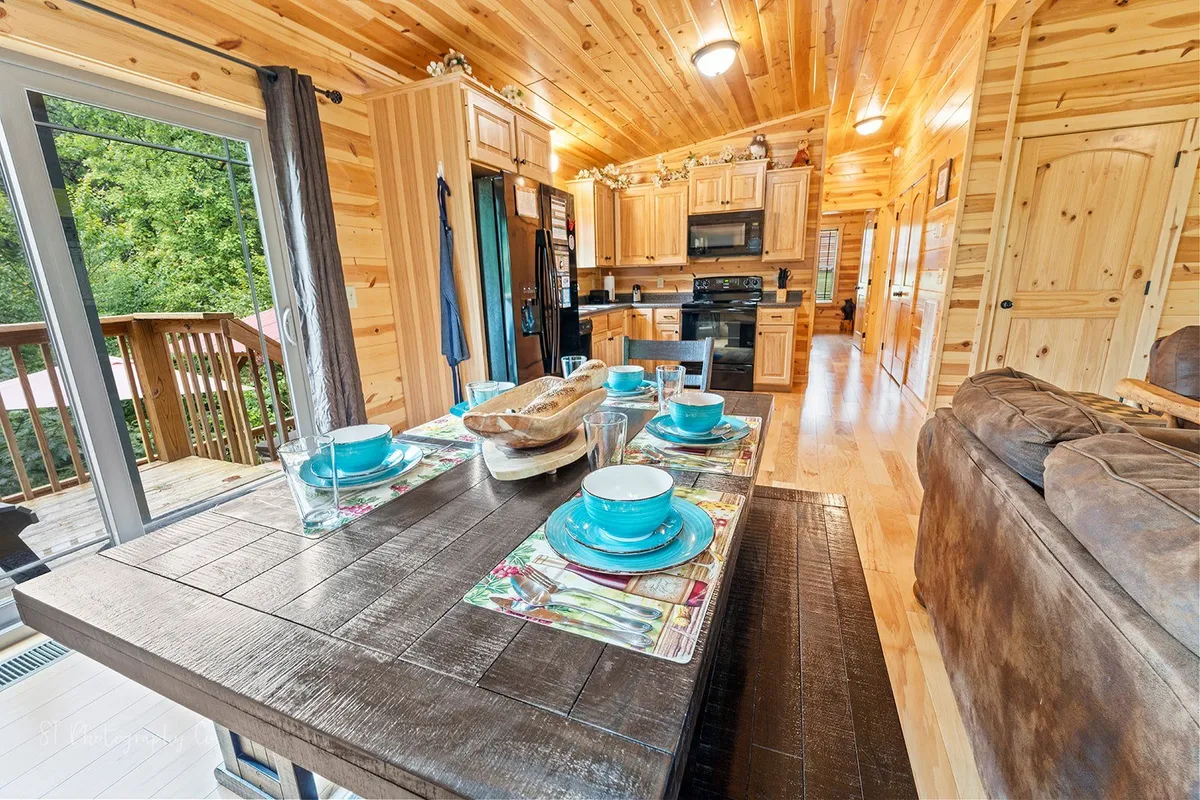A warm and inviting dining area featuring a dining table set with plates and glasses, kitchen cabinetry in the background, and an open doorway leading directly to the deck outside. Natural light streams in, hinting at the seamless connection between indoor comfort and outside adventure.
