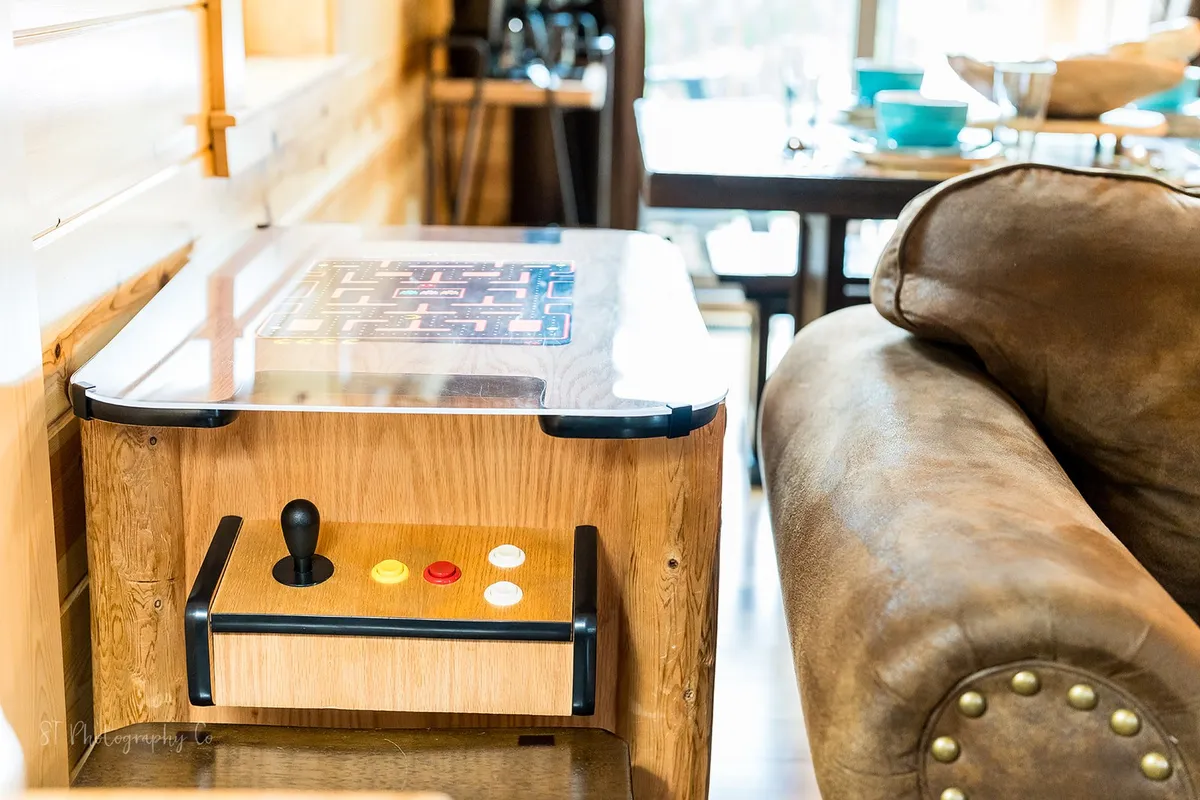 A comfortable living area featuring a brown couch, an arcade game table, and a dining table all clustered within the open lounge space. The setup encourages both relaxation and friendly competition, with the arcade game table positioned for easy access, and the couch inviting guests to gather, chat, or watch TV.