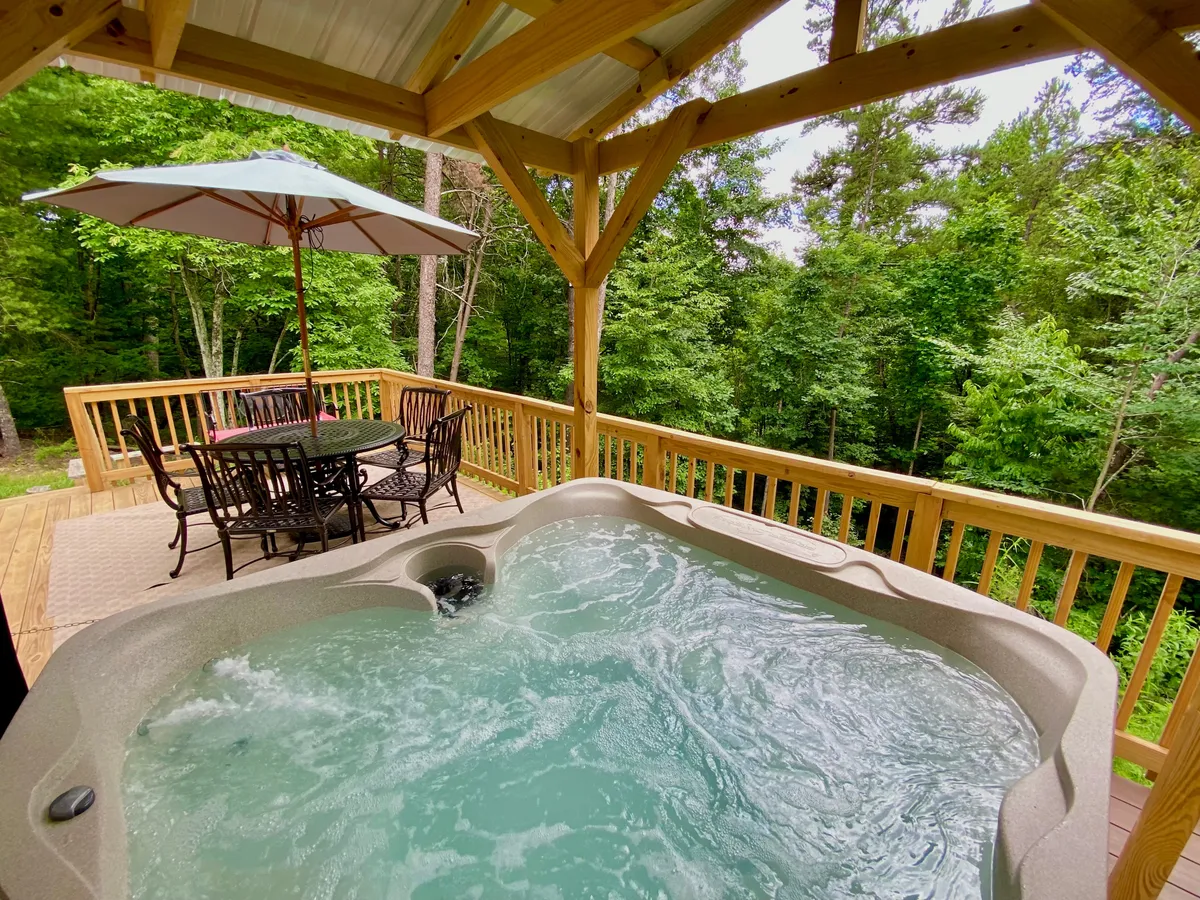 This exterior image shows a spacious wooden deck nestled among the trees, featuring a hot tub, a large dining table with an umbrella, wooden railing, and lush forest views. The setting evokes a tranquil retreat ideal for outdoor relaxation and gatherings, highlighting the connection to nature.