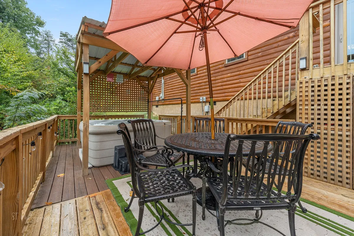 A spacious wooden deck featuring a dining table with umbrella, a hot tub, and views of surrounding greenery. The space is ideal for alfresco meals or relaxing soaks, surrounded by nature and fresh air.