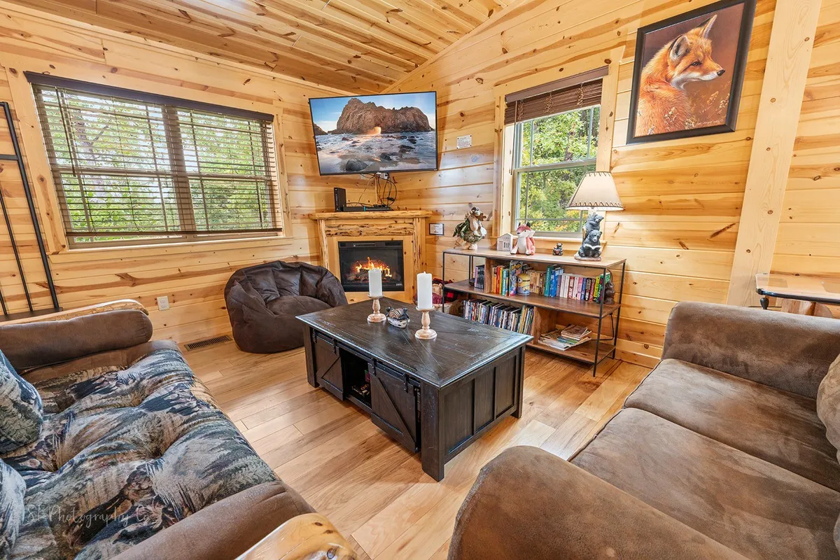 A warmly lit living area with a modern electric fireplace as the focal point, surrounded by comfortable sofas, a TV, and a bookshelf. The space is styled for relaxation, movie nights, and reading, with plenty of room for socializing.