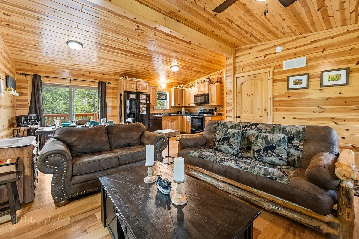 An open-concept space showing a comfortable sofa, coffee table, visible kitchen area, wooden beams, and large windows. The scene blends the kitchen, dining, and living areas, suggesting a seamless flow ideal for socializing, cooking, and relaxing together.