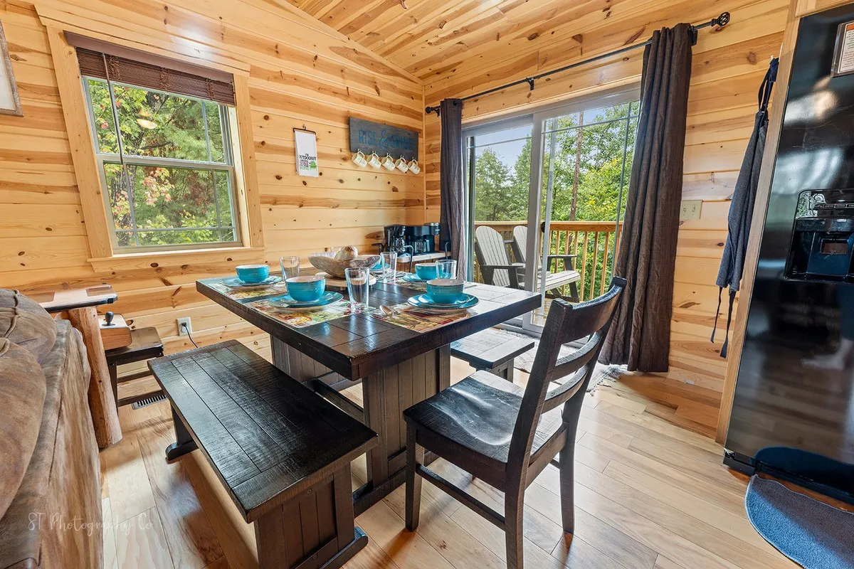 A cozy dining area with a wooden dining table set with blue dishes, a large window streaming in natural light, and wooden accents throughout. The setup evokes a sense of togetherness, with intimate seating for family meals or game nights, all bathed in welcoming daylight.