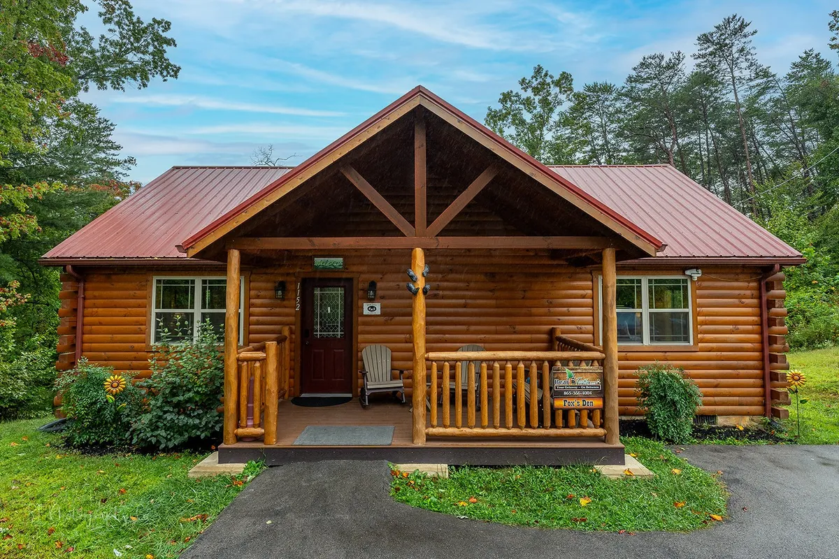 A cozy log cabin structure with a cheerful red metal roof and a welcoming front porch, complete with decorative chairs and signage. The exterior radiates warmth and embodies the classic Smoky Mountains getaway experience.