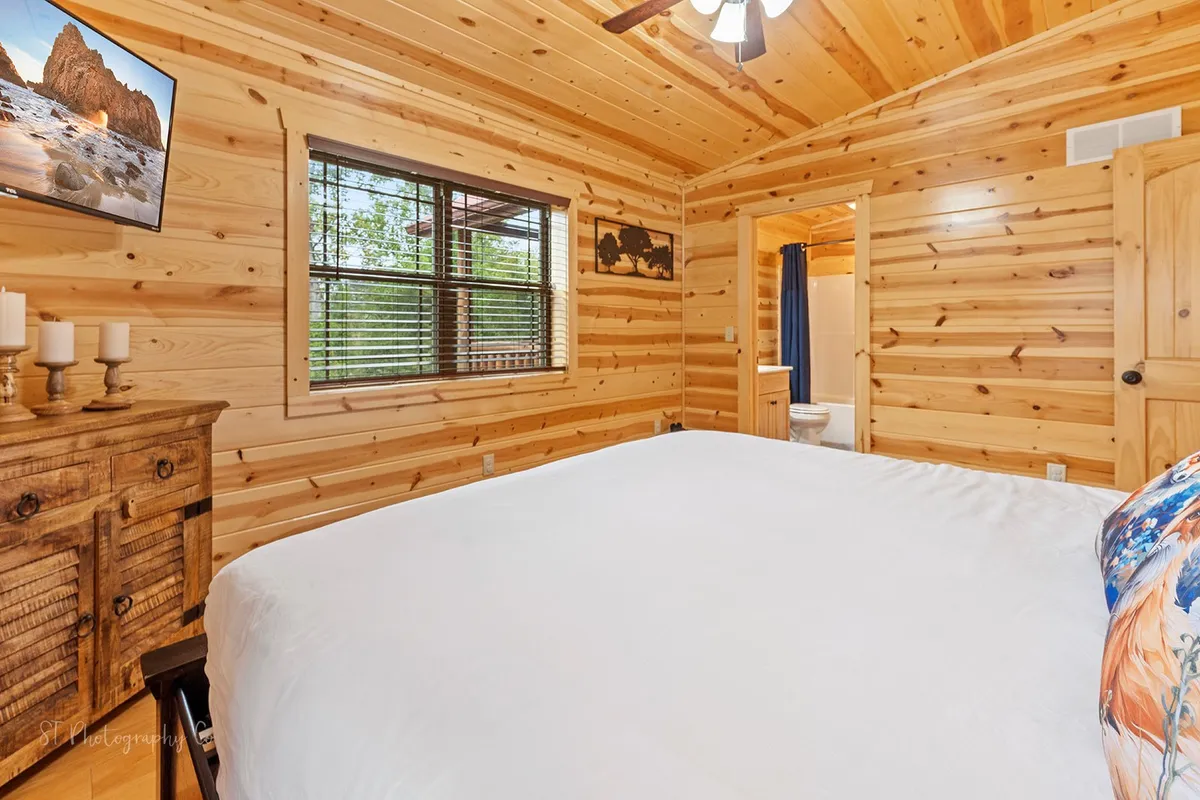 A spacious master bedroom featuring a large bed with crisp white bedding, rustic wooden furniture, and a large window offering serene views of the outdoors. The framed landscape photograph above the bed and warm wood accents create a tranquil and comfortable atmosphere.